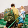 boy wears his state bags kane kids double pocket large backpack in olive diagonal zipper with his friends on green bleachers.